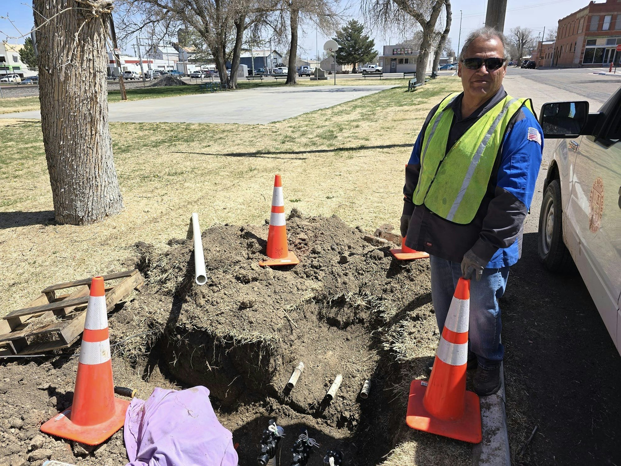 A worker with traffic cones by an excavation site near a tree and vehicle.
