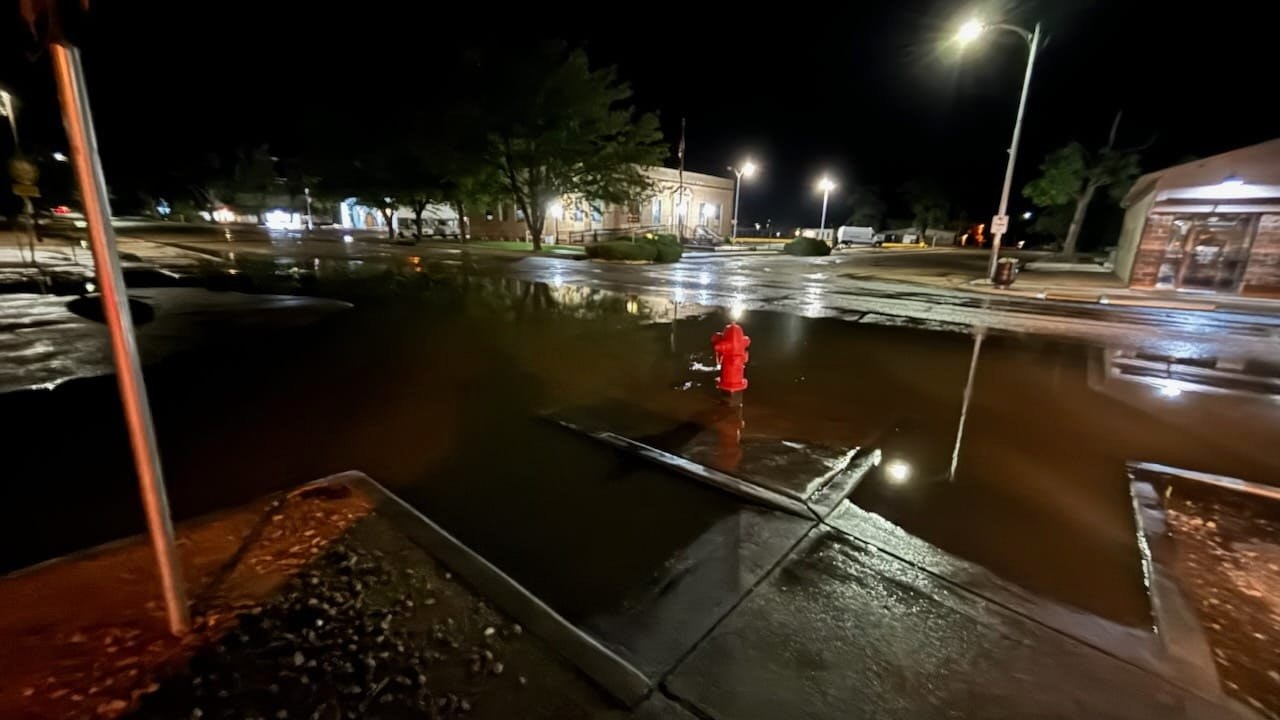 A flooded street at night with a red fire hydrant visible and reflections of lights on the water.