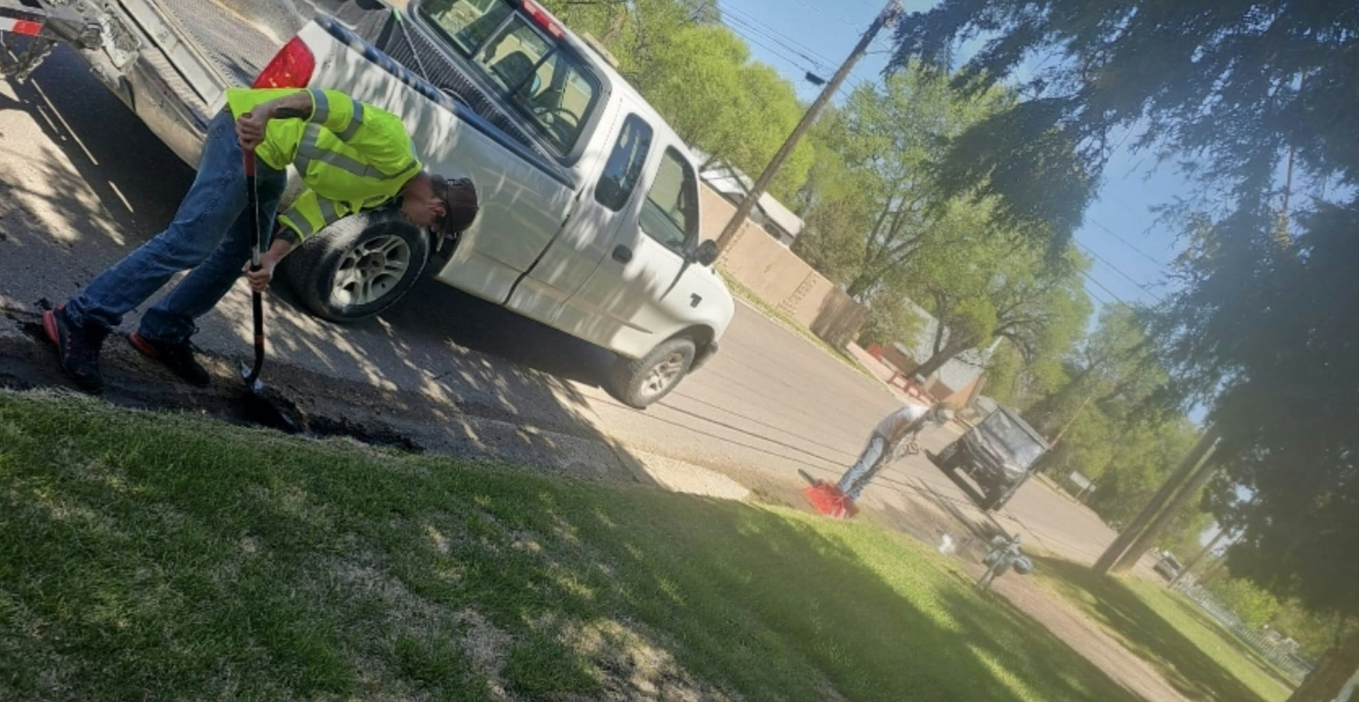 Two people working on a street, one with a shovel by a pickup truck, in a sunny, tree-lined area.