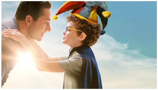 A man smiles at a child in a jester hat and cape, against a sunny sky.