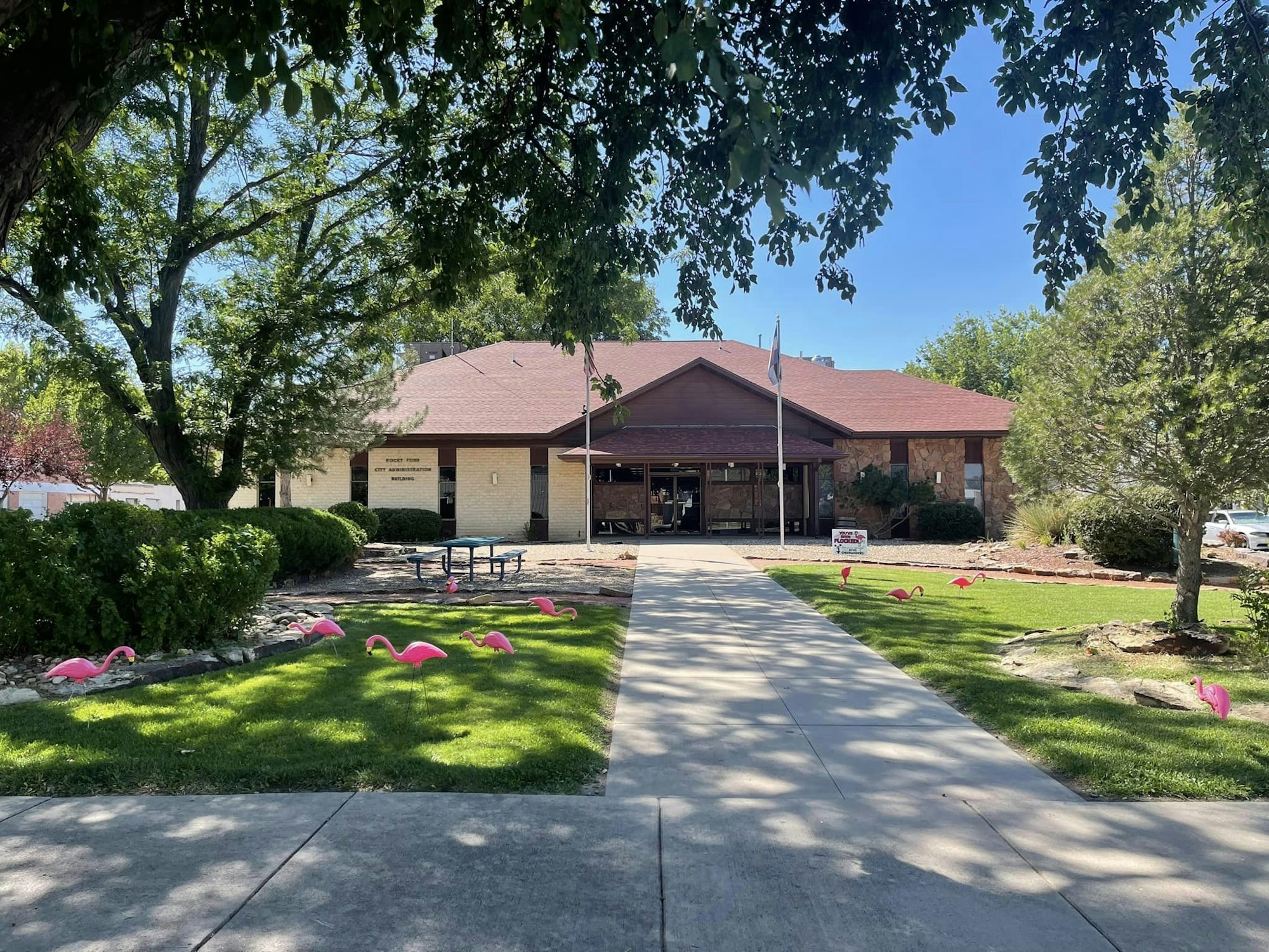 Building with a sign, pink flamingo decorations on the lawn, trees, and a paved walkway.