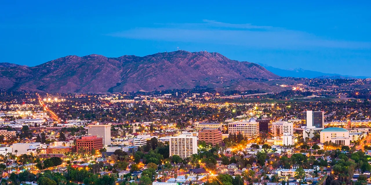 Cityscape at dusk with lights, buildings, and mountains in the background.