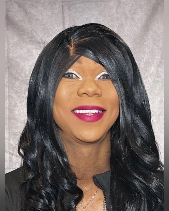 A person with black hair, bright lipstick, and a wide smile against a grey backdrop.