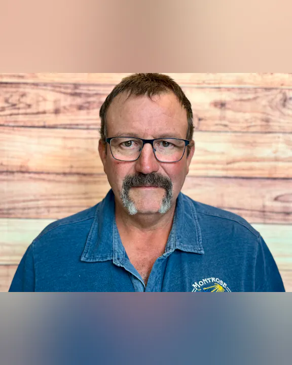 A middle-aged man with glasses and a mustache, wearing a blue shirt, stands in front of a wooden background.