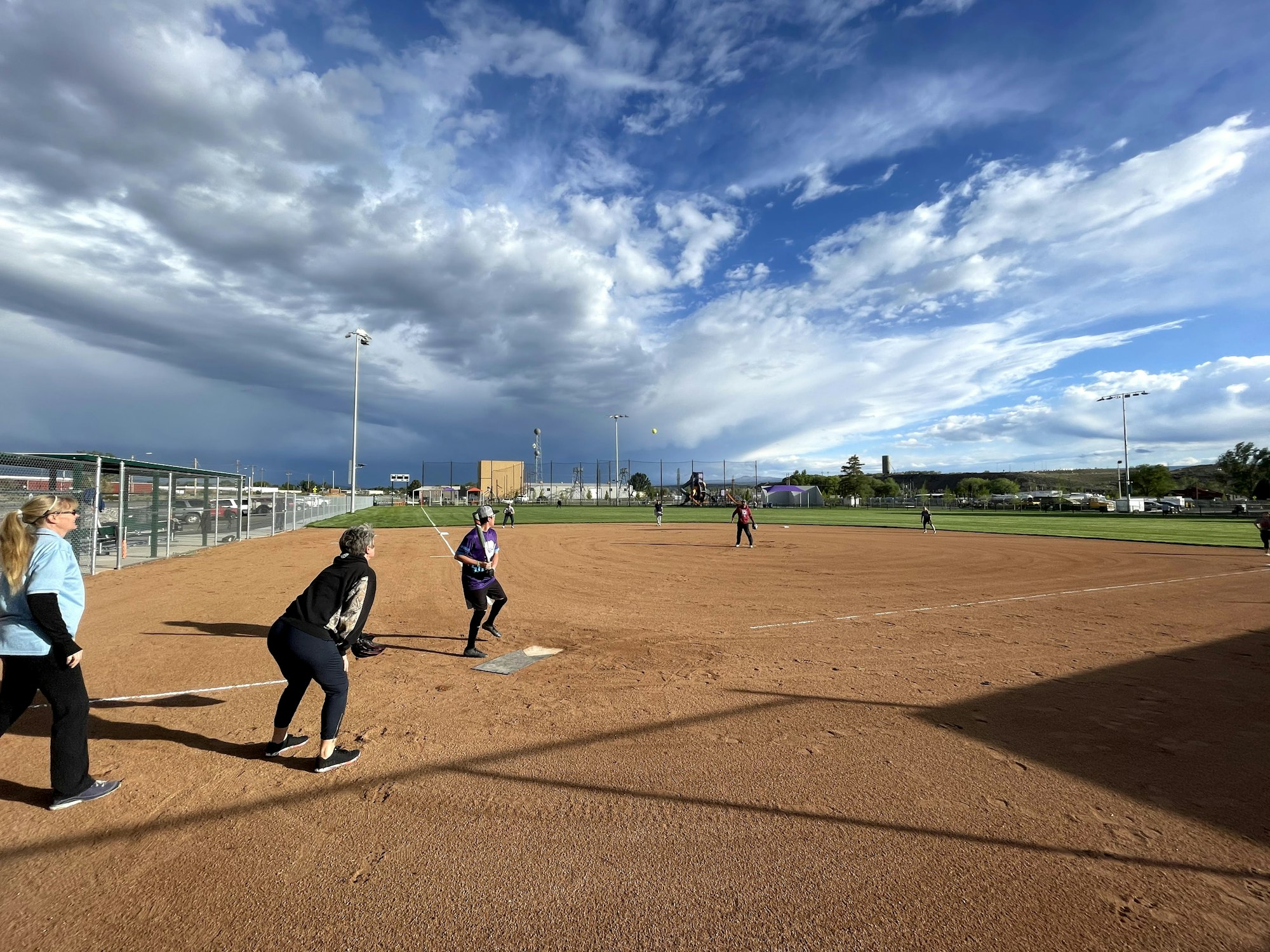 A softball game with players on the field, dramatic clouds overhead, casting long shadows on the ground.