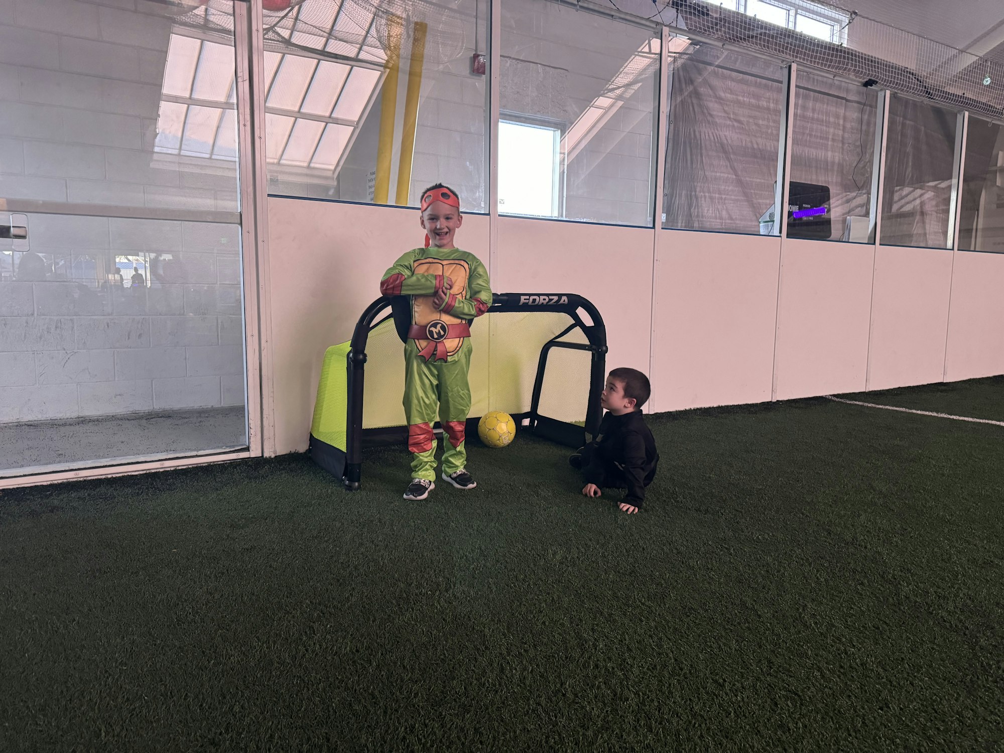 Two kids playing in an indoor area with artificial turf. One is in costume, standing by a mini goal; the other is on the ground by the ball.
