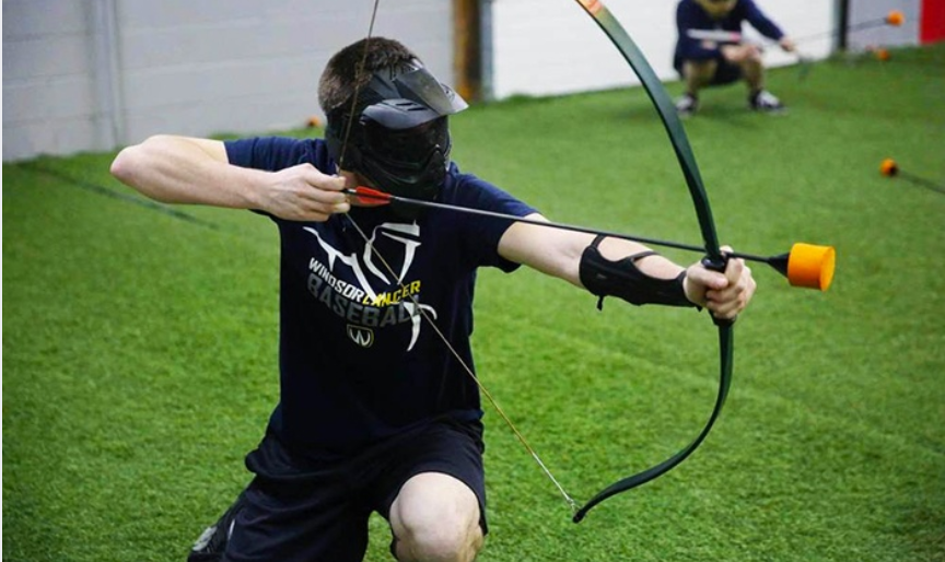 A person crouches aiming a bow with a foam-tipped arrow in an indoor setting, wearing a face mask and sports gear.