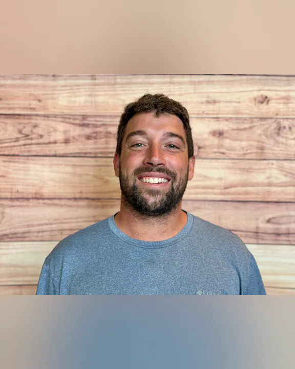 The image shows a man smiling in front of a wooden wall backdrop, wearing a gray t-shirt.