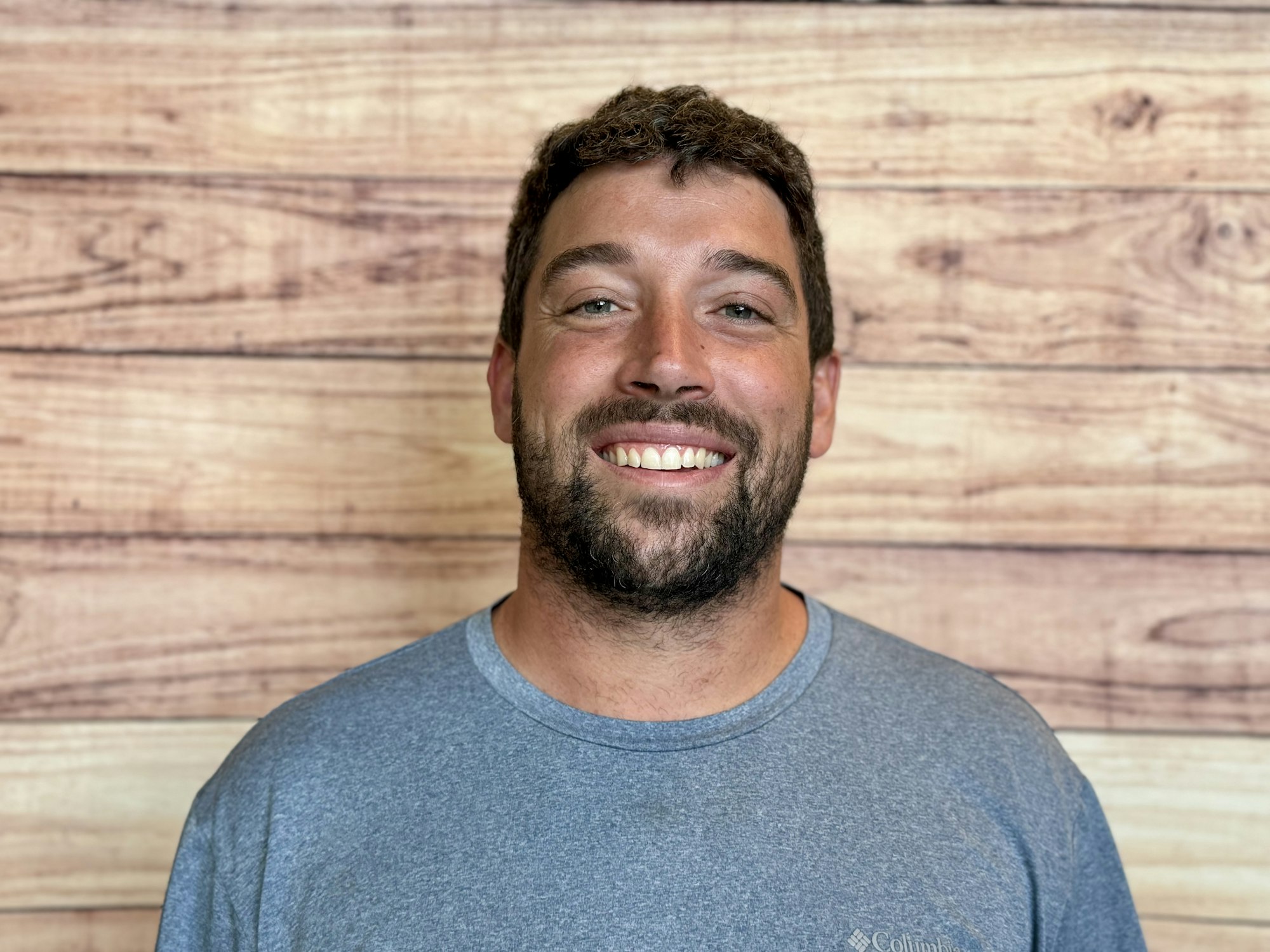 The image shows a man smiling in front of a wooden wall backdrop, wearing a gray t-shirt.