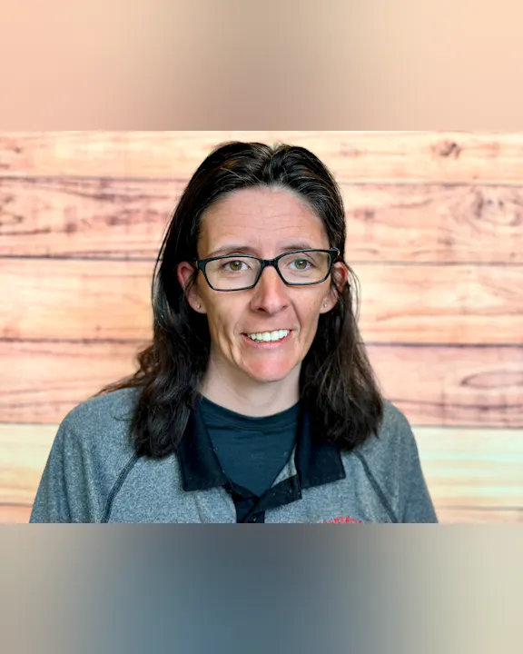 A person with long dark hair and glasses smiles, wearing a grey top, against a wooden backdrop.
