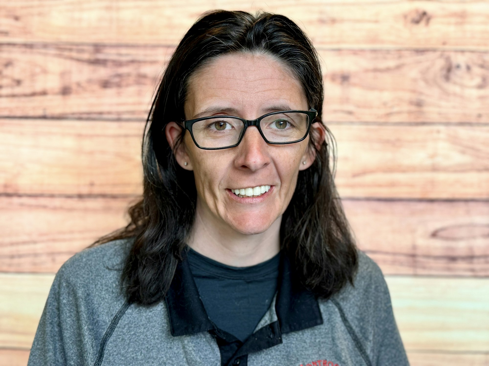 A person with long dark hair and glasses smiles, wearing a grey top, against a wooden backdrop.