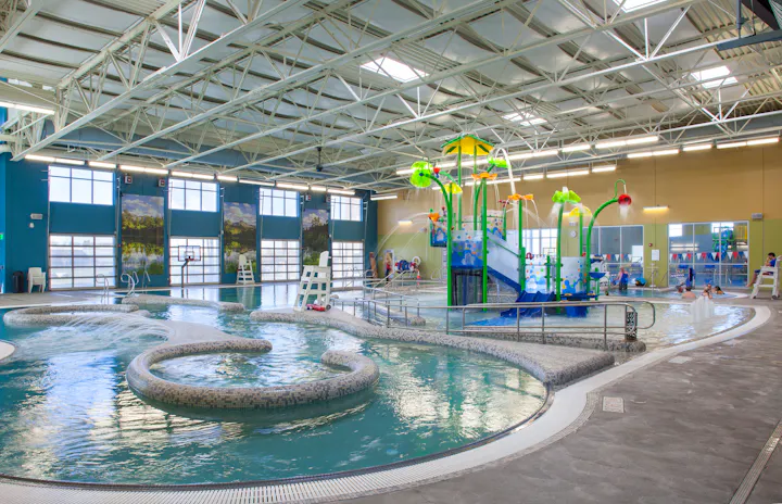 An indoor swimming pool featuring a water slide, play structures, and areas for relaxation. Families enjoying the space.