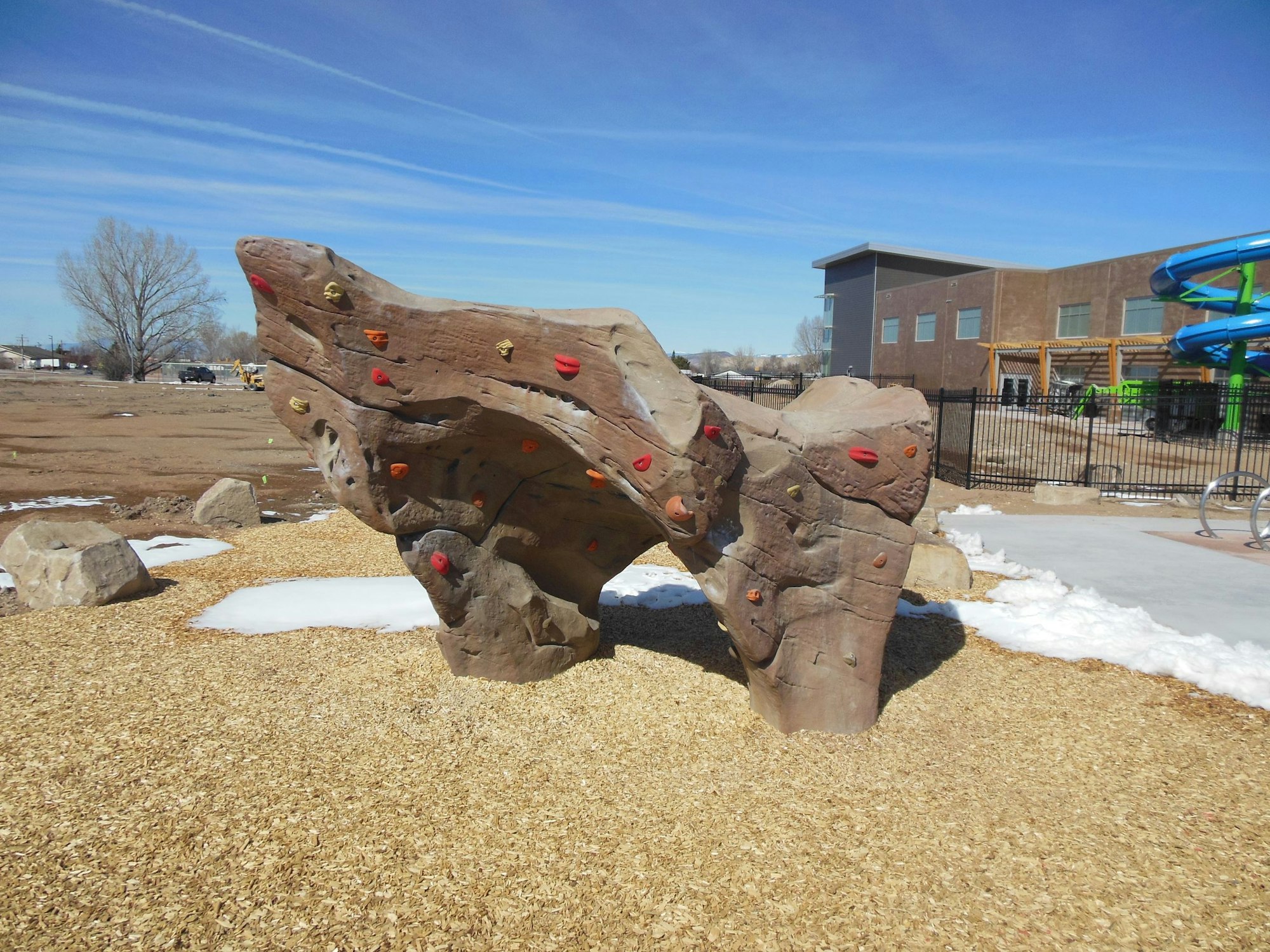 A climbing boulder with colored holds, surrounded by wood chips and rocks, near a building and a water slide on a sunny day.