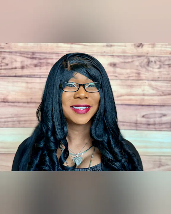 The image shows a smiling person with long, black, wavy hair, wearing glasses and necklaces, against a wooden background.