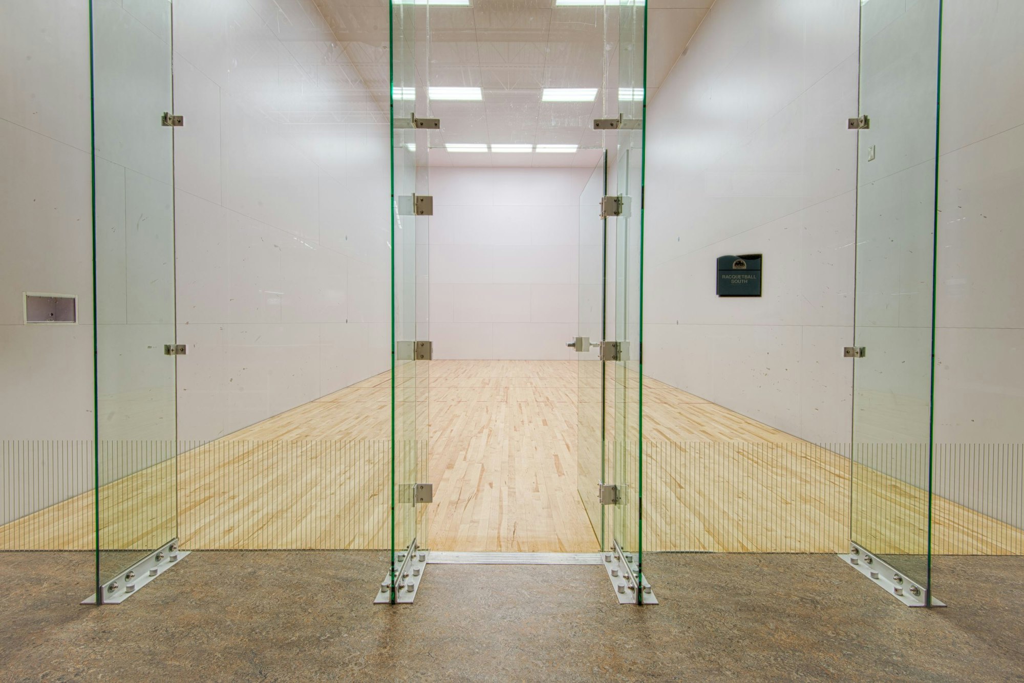 The image shows the entrance to a racquetball court with glass doors and a wooden floor inside.