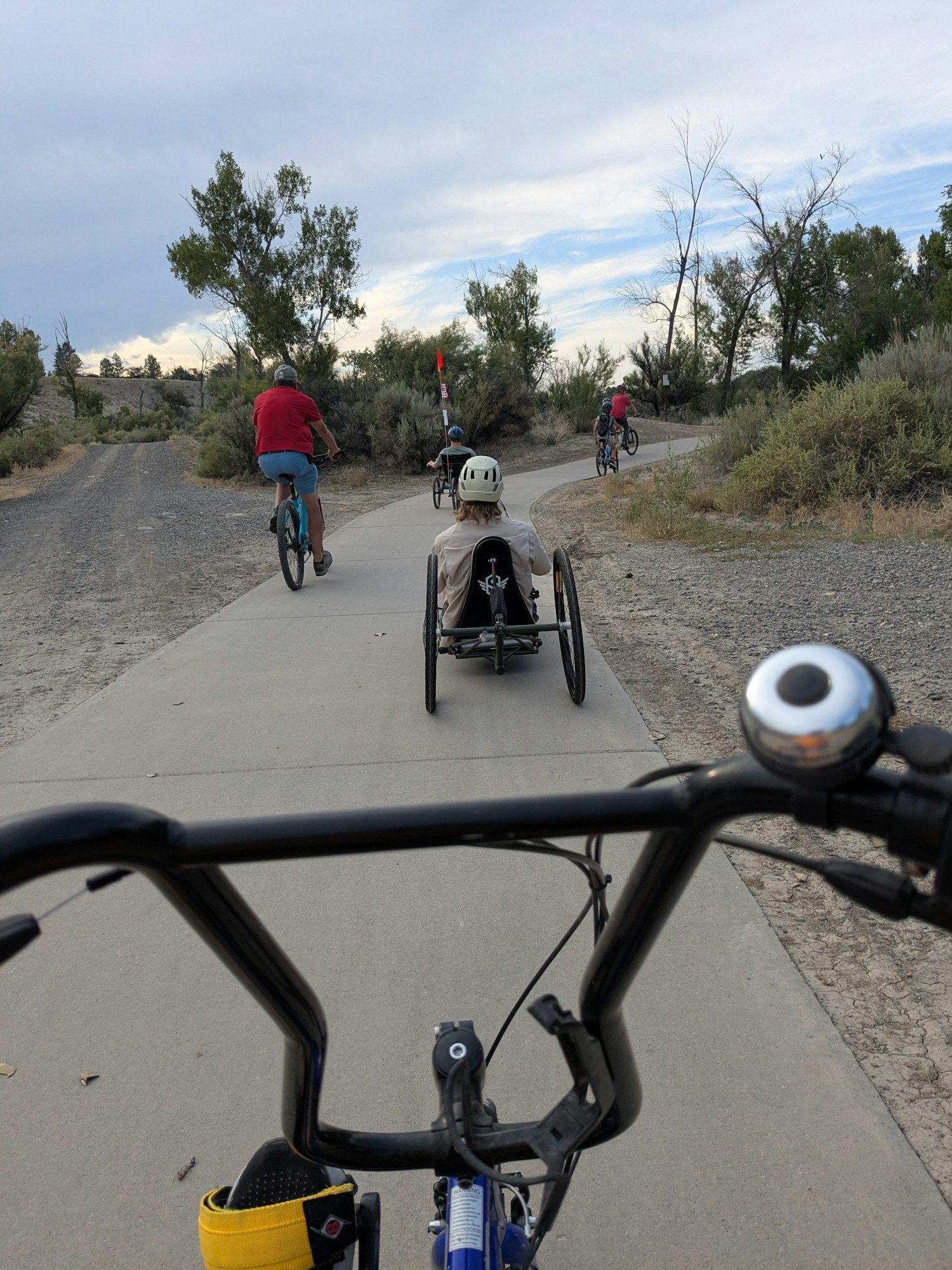 A group of cyclists, including some using adaptive bikes, riding on a paved path through a natural setting.