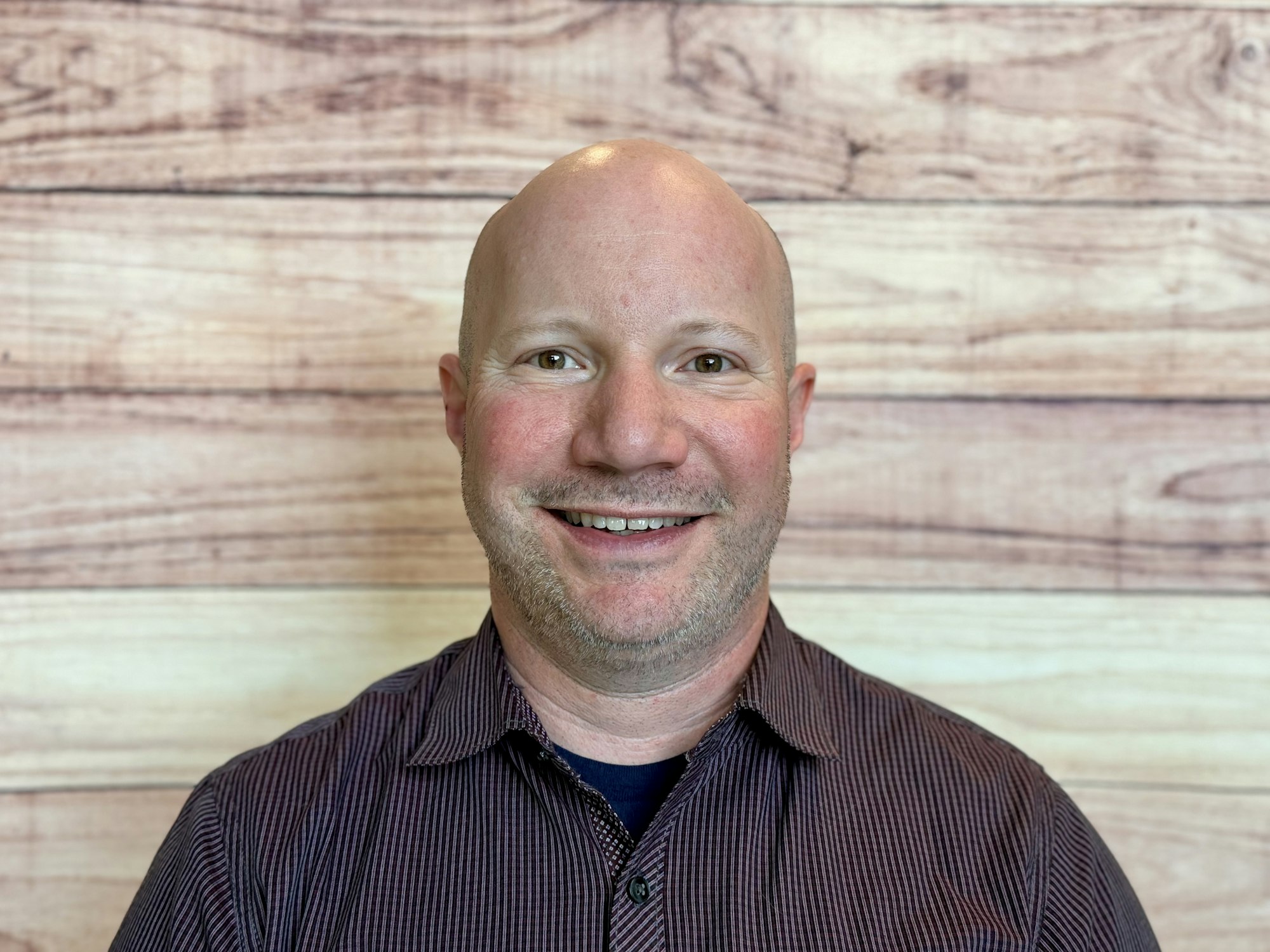 A smiling bald man in a checkered shirt poses in front of a wooden background.