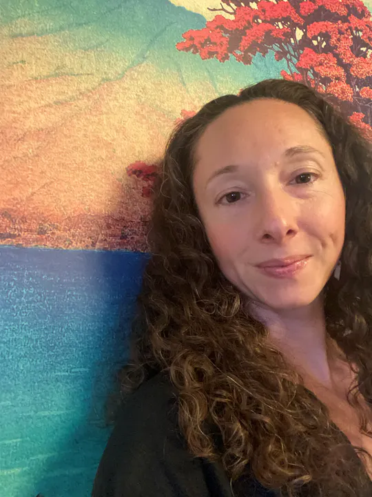 A woman with curly hair smiles in front of a colorful landscape mural featuring blue water and red trees.