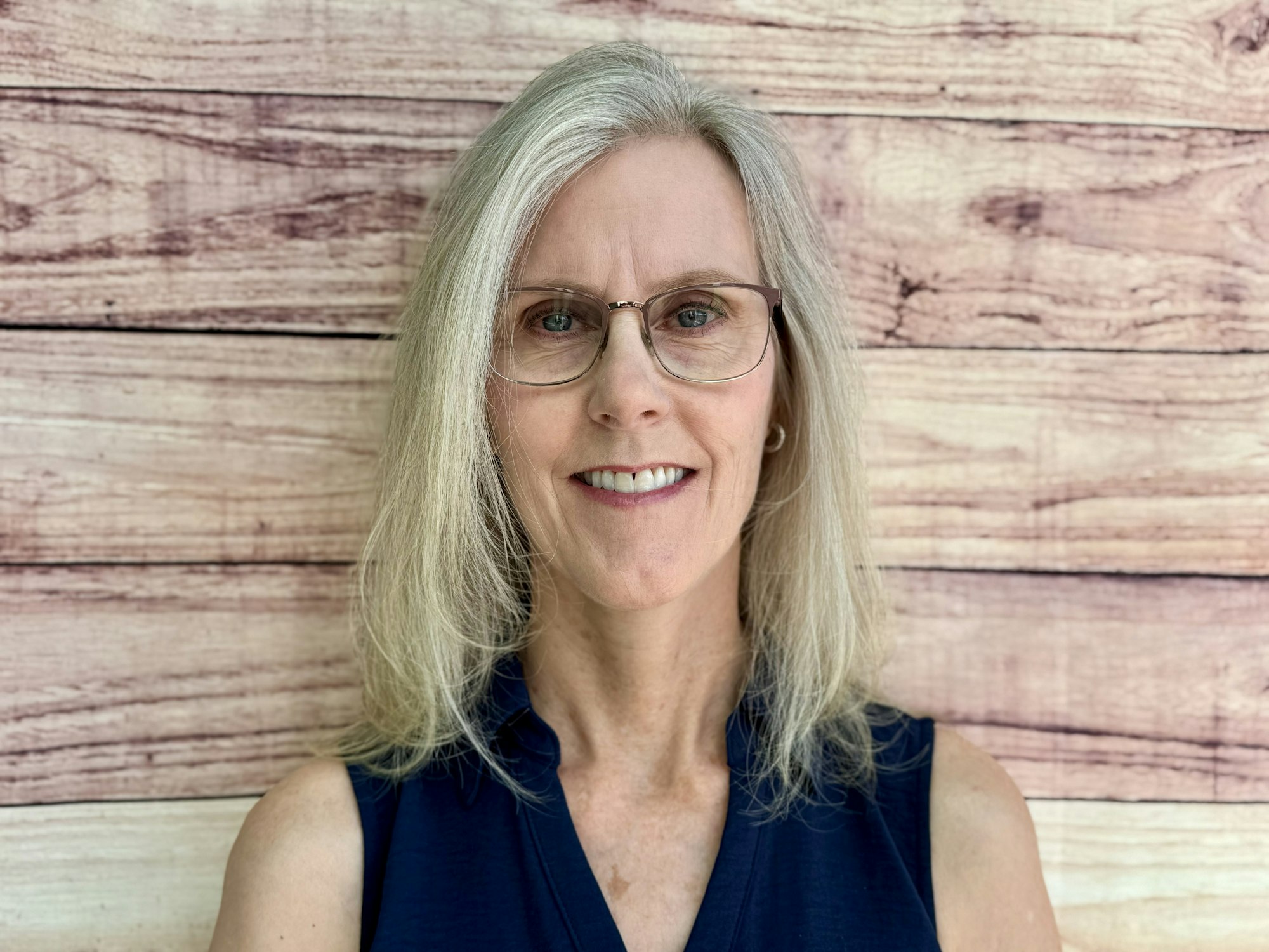 A woman with light blonde hair and glasses smiles against a wooden background. She's wearing a dark sleeveless top.
