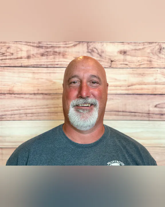 A smiling man with white facial hair and a bald head, wearing a gray tee shirt, posing against a wood-patterned background.