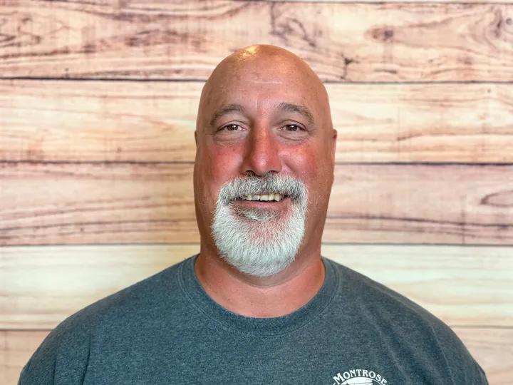 A smiling man with white facial hair and a bald head, wearing a gray tee shirt, posing against a wood-patterned background.