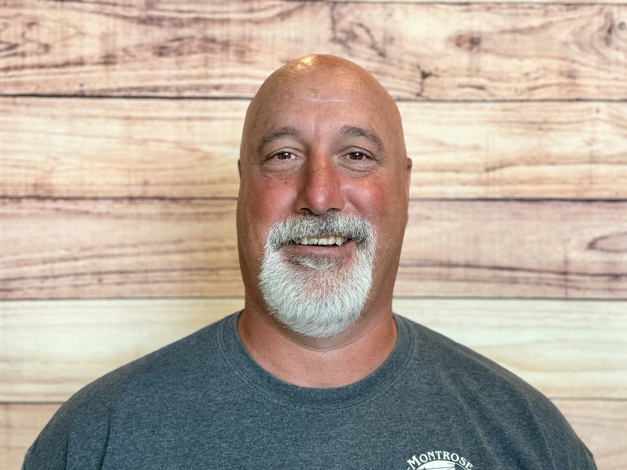 A smiling man with white facial hair and a bald head, wearing a gray tee shirt, posing against a wood-patterned background.