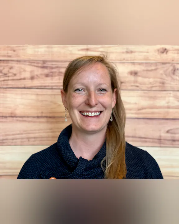 A smiling individual with long blonde hair, wearing a dark sweater, is in front of a wooden backdrop.
