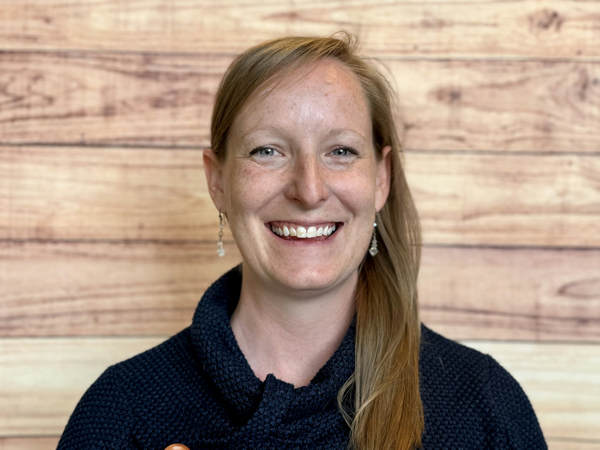 A smiling individual with long blonde hair, wearing a dark sweater, is in front of a wooden backdrop.