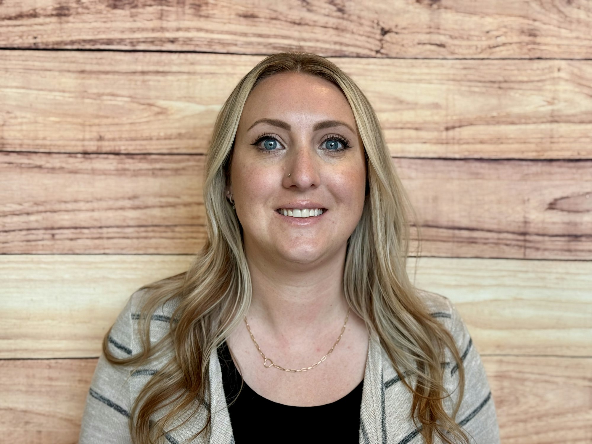 A woman with long blonde hair smiles against a wooden background, dressed casually in a black top and a striped cardigan.