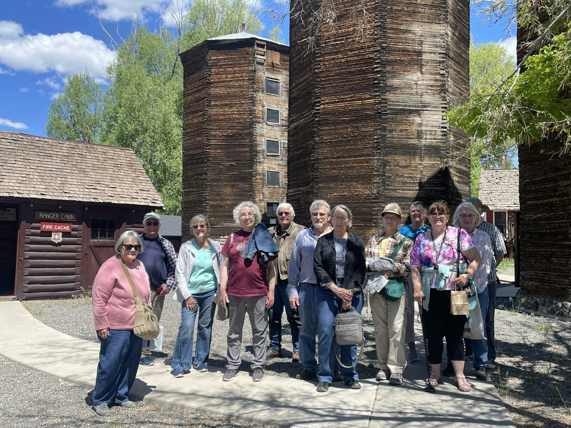 A group of people posing in front of rustic buildings and silos outdoors on a sunny day.