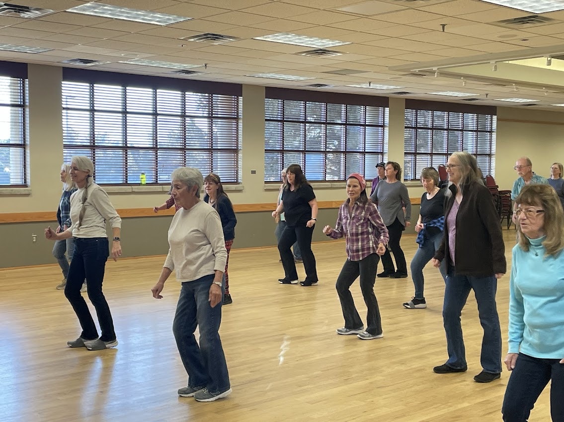 A group of people line dancing in a large room with wooden floors and bright windows.