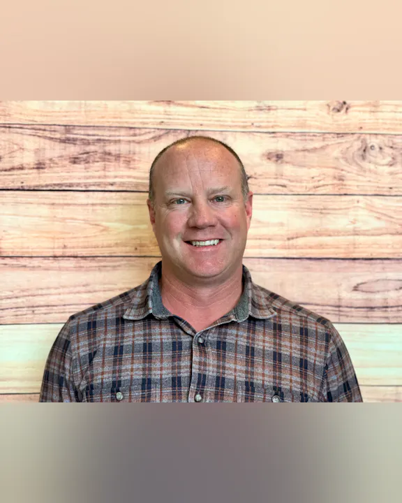 A smiling man wearing a plaid shirt, standing in front of a wooden background.