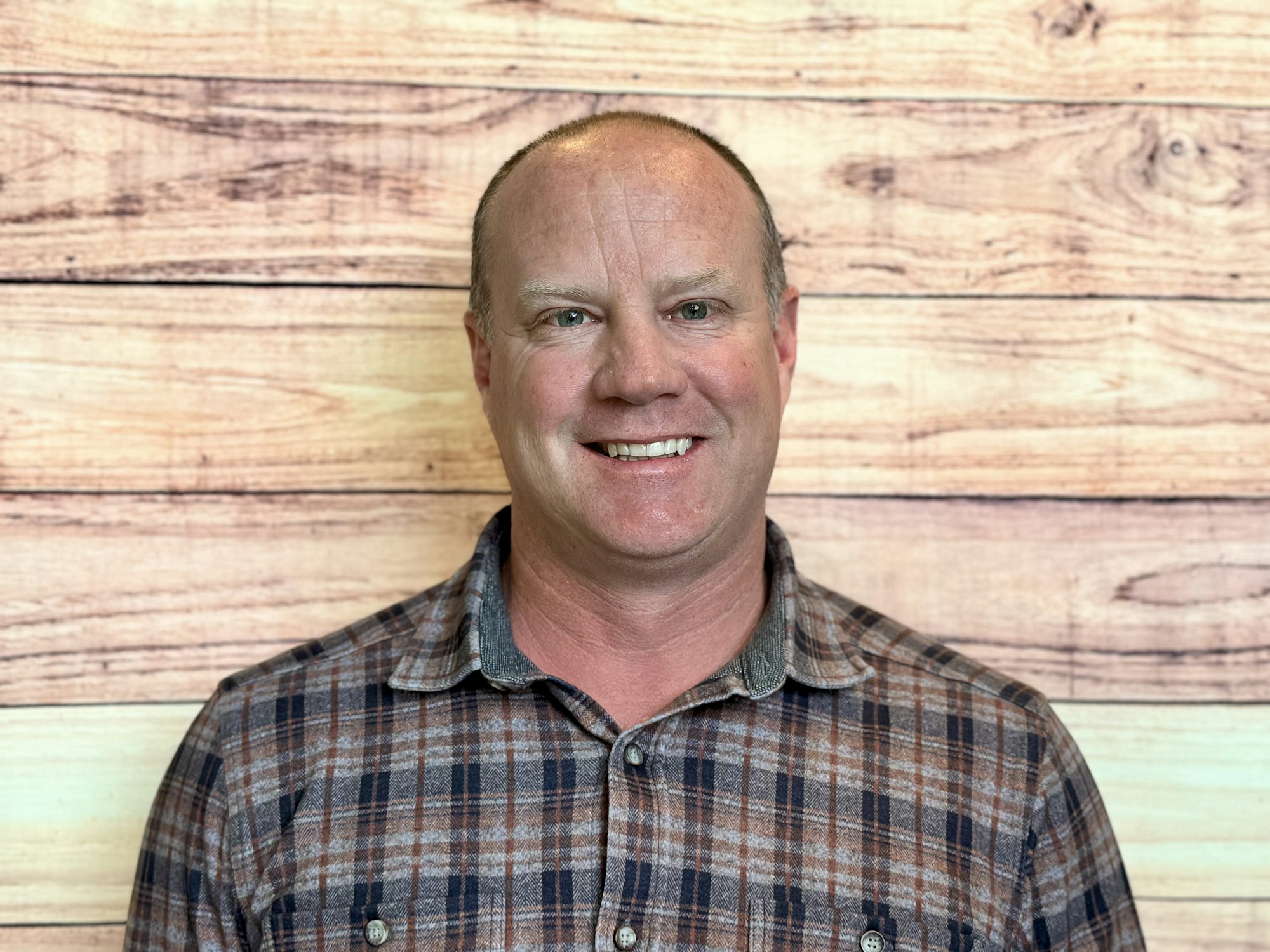 A smiling man wearing a plaid shirt, standing in front of a wooden background.