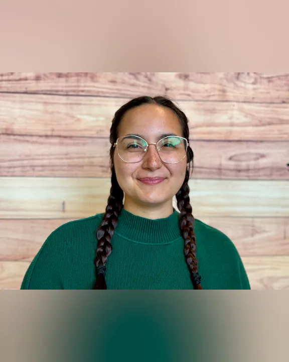 A smiling person with long braids, wearing glasses and a green sweater, stands in front of a wooden background.
