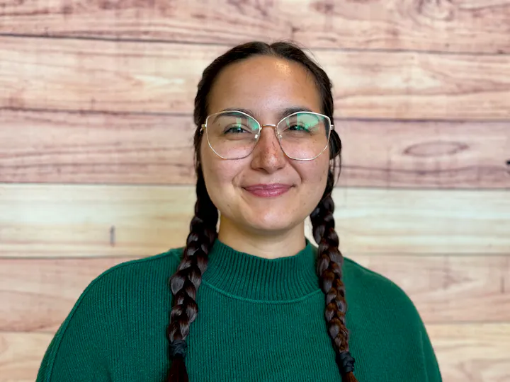 A smiling person with long braids, wearing glasses and a green sweater, stands in front of a wooden background.