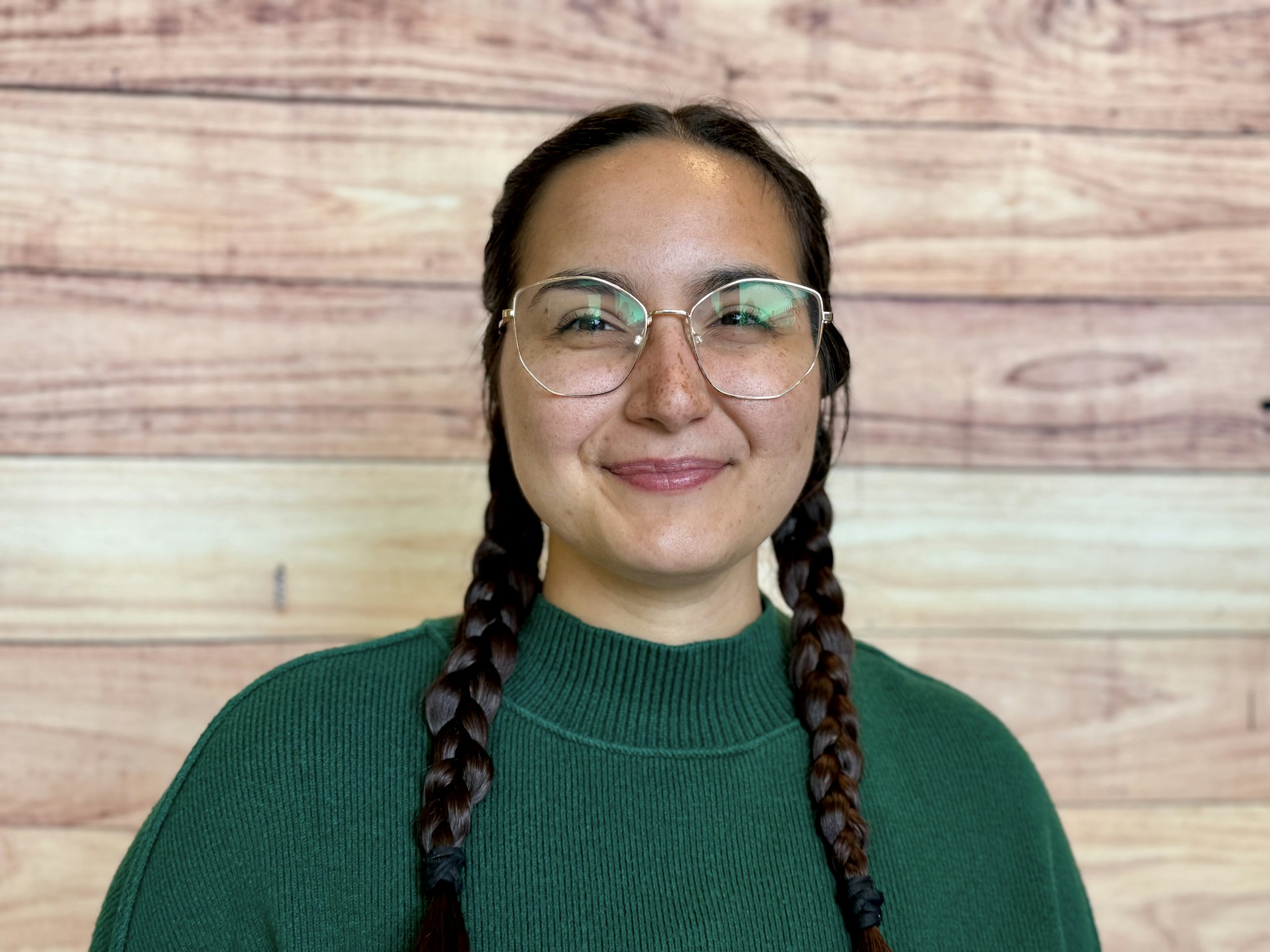 A smiling person with long braids, wearing glasses and a green sweater, stands in front of a wooden background.