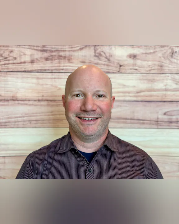 A smiling person with a bald head wearing a plaid shirt, set against a wooden backdrop.