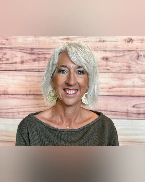 A smiling woman with short, light hair wearing large earrings, posing against a wooden background.