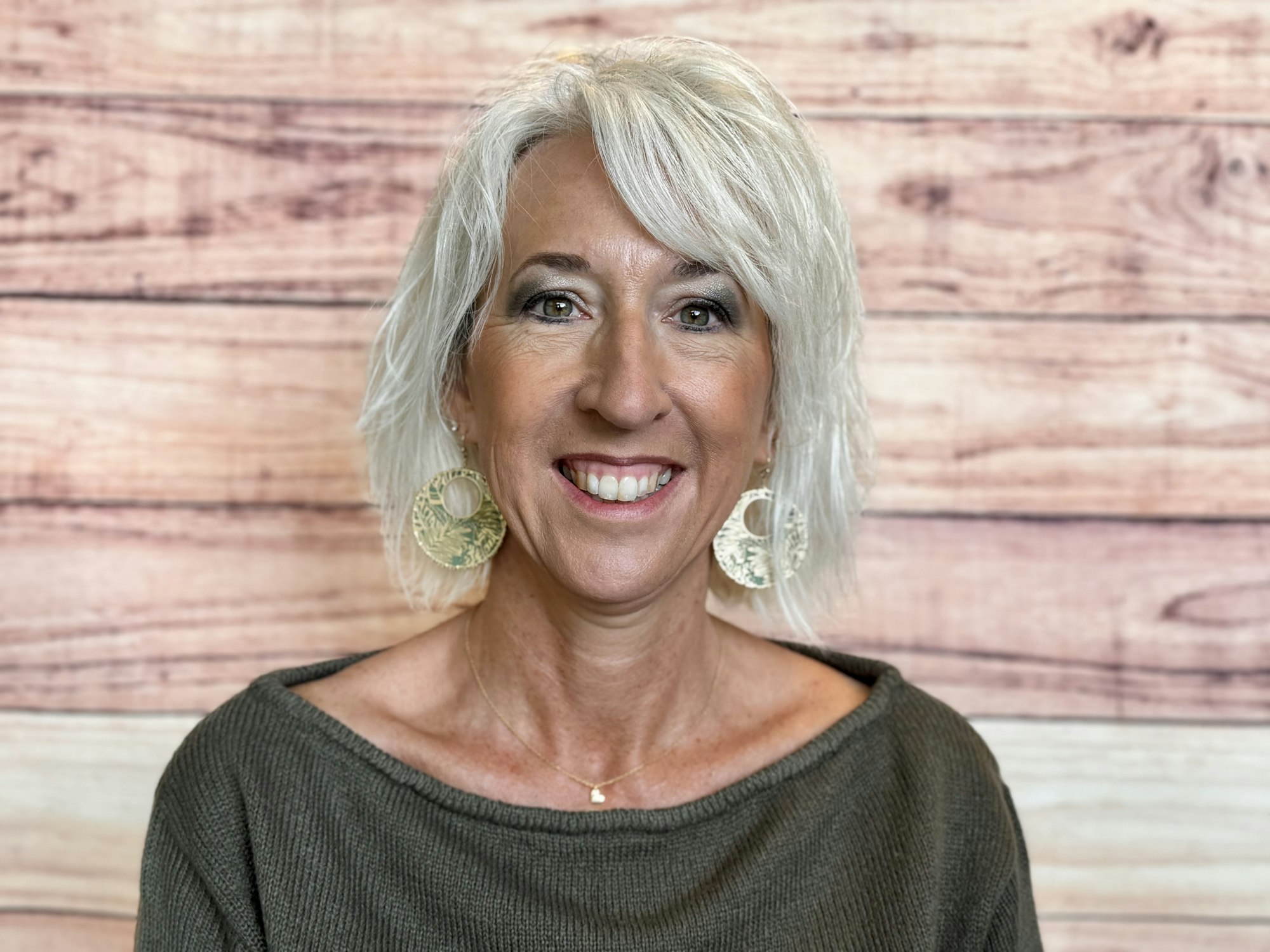 A smiling woman with short, light hair wearing large earrings, posing against a wooden background.