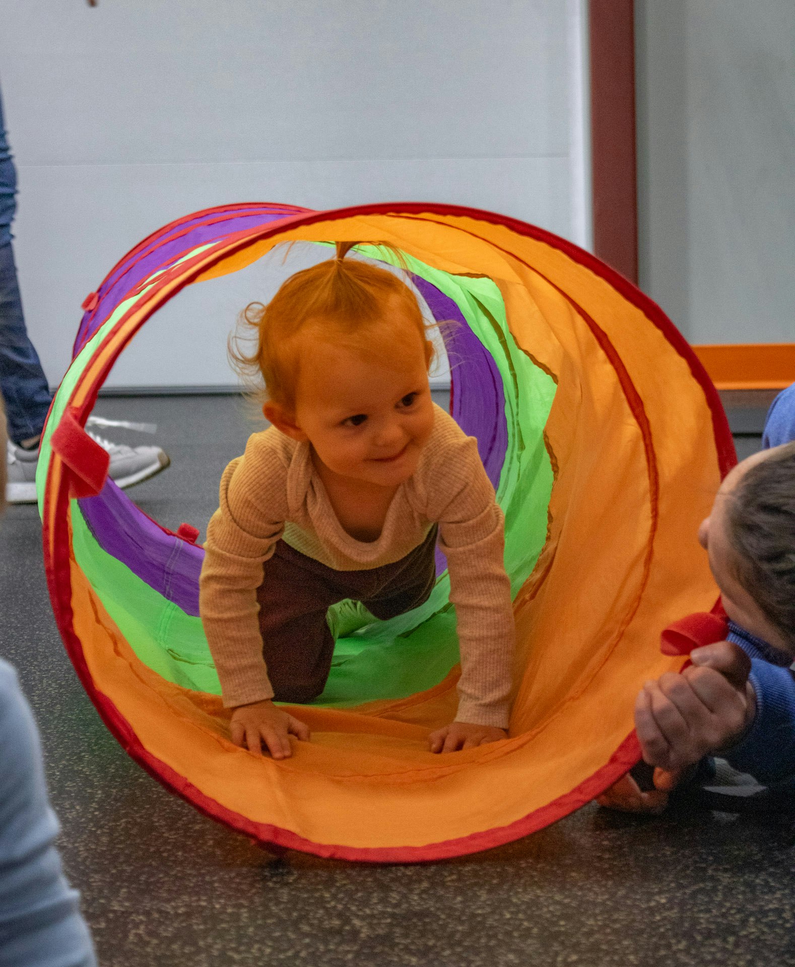 A smiling infant crawls through a colorful play tunnel, with adults nearby at Flex Rec.
