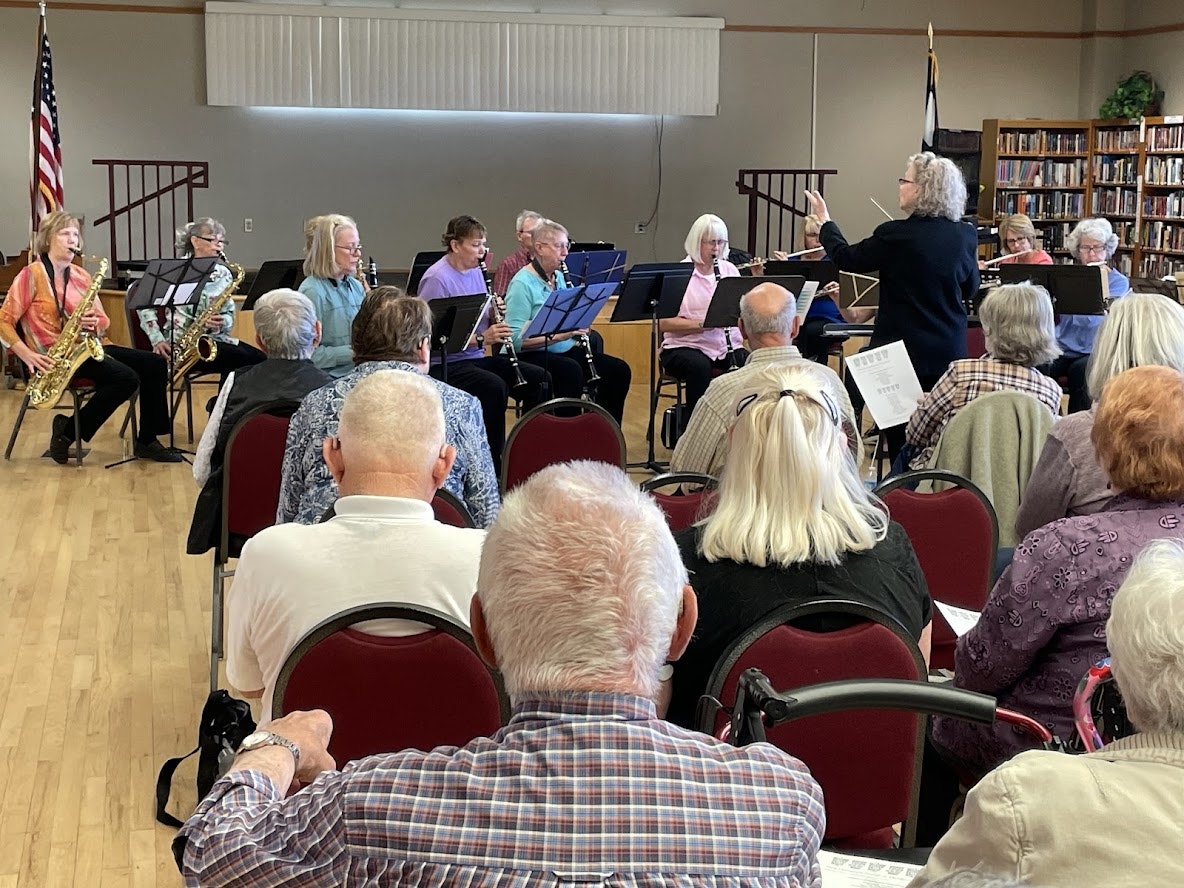 A music ensemble with people playing instruments, led by a conductor, performing for an audience in a indoor setting.