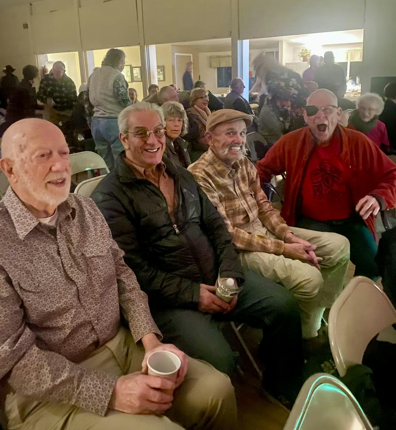 A group of older adults sitting on chairs at an indoor event, smiling and holding cups.