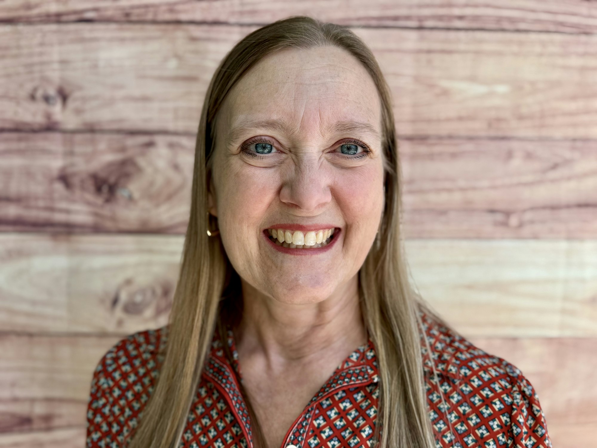 A smiling woman with long hair is posed in front of a wooden backdrop, wearing a patterned top.