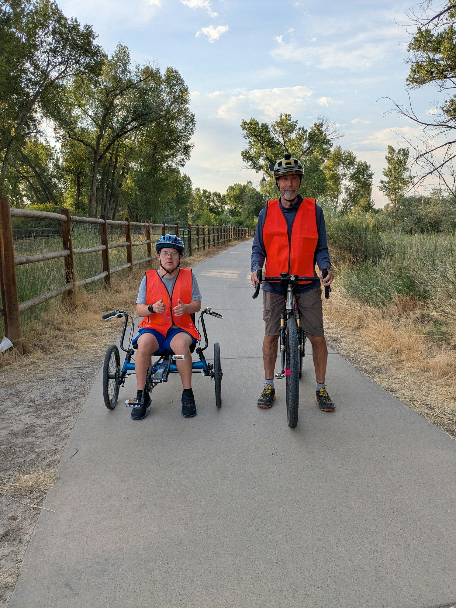 A boy and his father pose for a picture on a paved trail during an adaptive biking outing.ath surrounded by trees.