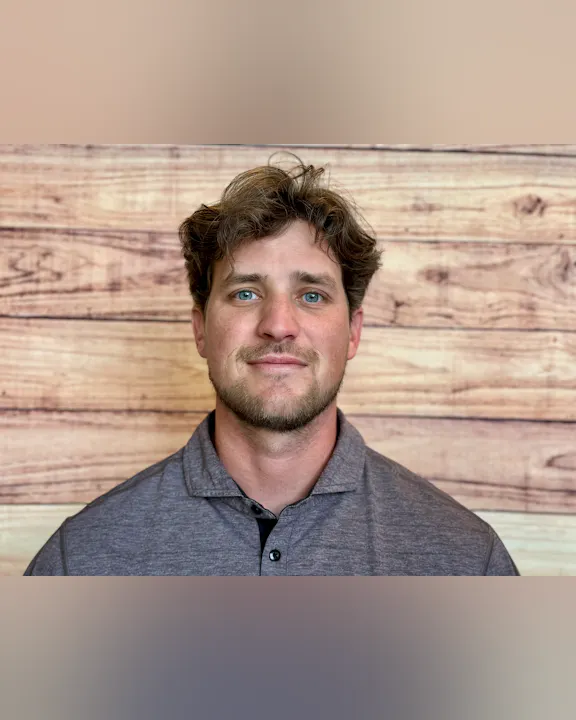 A man with light brown hair and blue eyes is smiling, wearing a gray polo shirt, against a wooden background.
