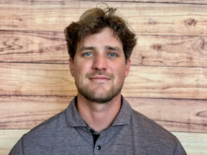 A man with light brown hair and blue eyes is smiling, wearing a gray polo shirt, against a wooden background.