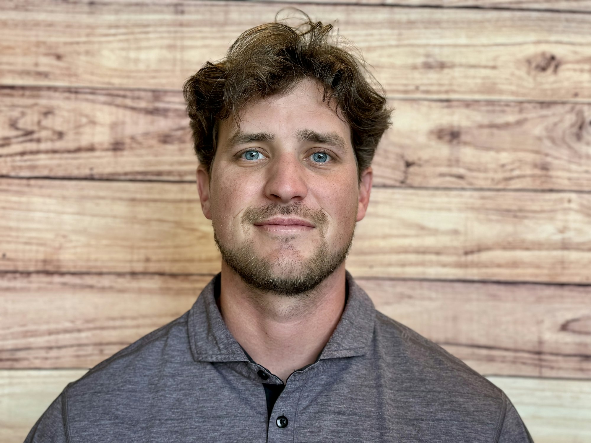 A man with light brown hair and blue eyes is smiling, wearing a gray polo shirt, against a wooden background.