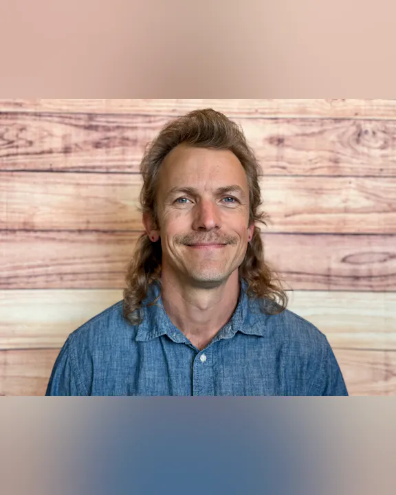 A smiling individual with a mustache and long hair is wearing a denim shirt, set against a wooden backdrop.