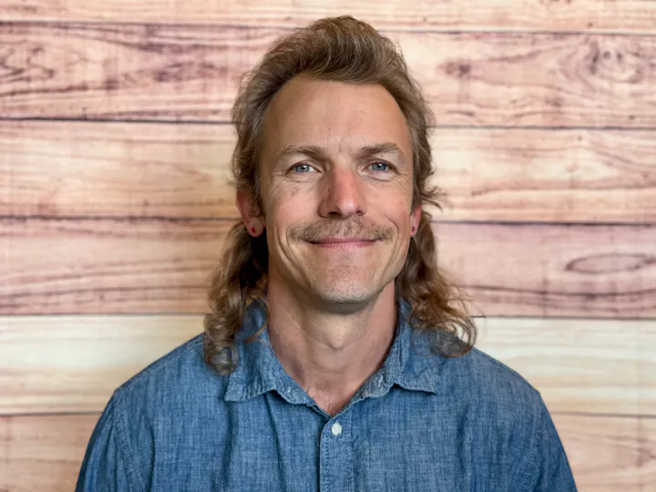 A smiling individual with a mustache and long hair is wearing a denim shirt, set against a wooden backdrop.