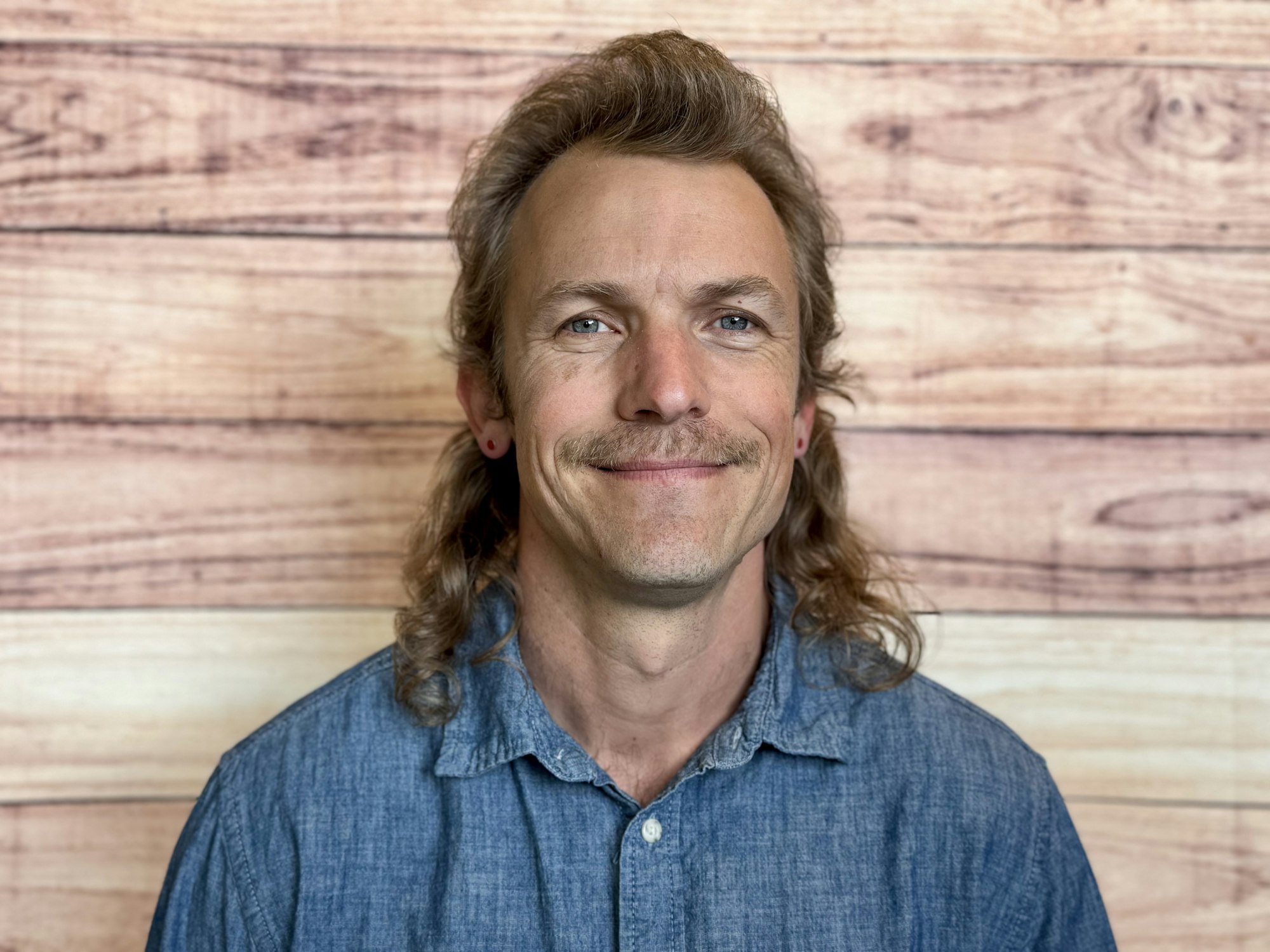 A smiling individual with a mustache and long hair is wearing a denim shirt, set against a wooden backdrop.